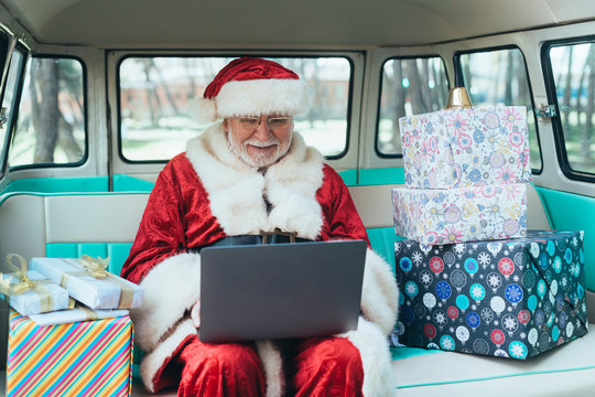 Cheerful man in costume of Santa Claus sitting in van with laptop and colorful gifts on sunny day