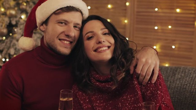 Happy Smiling Couple Holding Glasses of Champagne Rejoicing Sitting near Christmas Tree at Warm and Cozy Home Atmosphere Holding Glasses of Chamagne Looking to Camera Concept of Holidays and New Year.