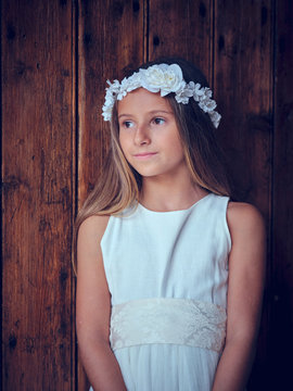 Cute little girl in white long dress and headband resting leaning on wooden door with ivy looking away