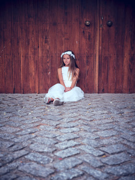 Cute little girl in white long dress and headband resting sitting near wooden door with ivy looking away