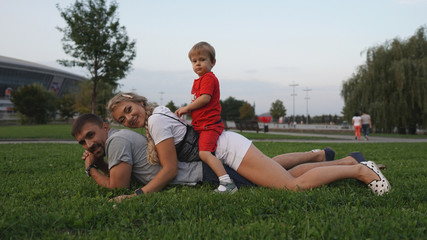 Naklejka na ściany i meble Happy family - dad, mom and little son lying on grass in the park in summer.