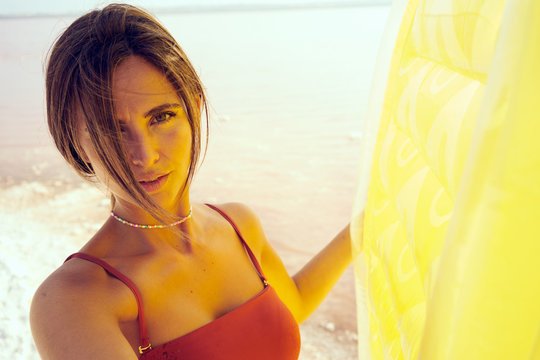 Serene woman in red swimwear with bright air mattress standing on seashore in sunlight looking at camera