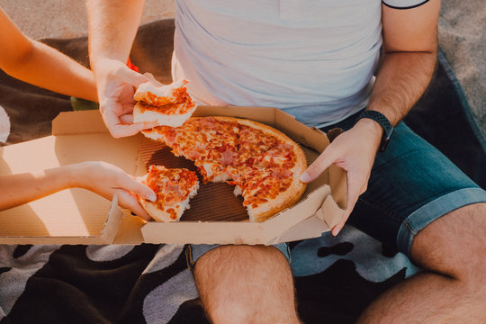 Crop and unrecognizable couple sitting on towel beach on sand eating pizza
