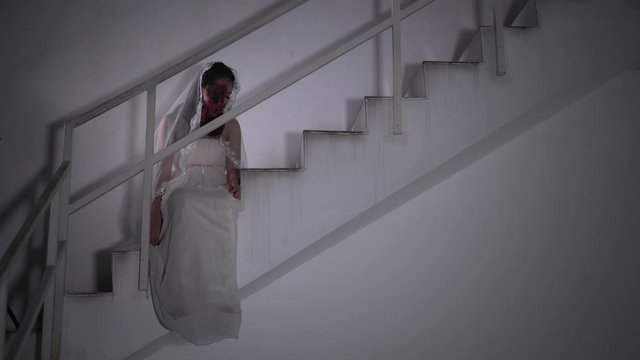 Scary female zombie or ghost with wounded face sitting on the stairs while wearing bride gown at home. Halloween concept. Shot in 4k resolution