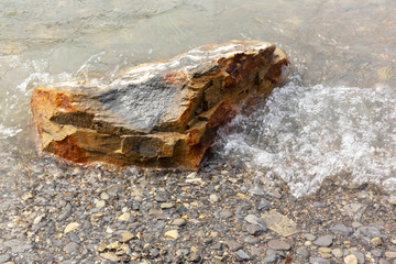 A brown, angular stone lies on the surf line half in the water.