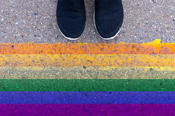 Cropped humans legs in black shoes standing on asphalt in front of lgbt rainbow colored flag,...