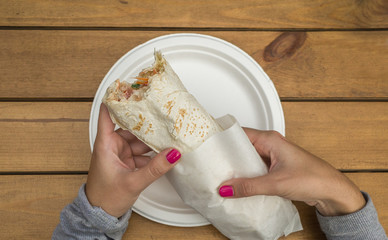 bitten doner kebab in female hands over wooden table, flat lay