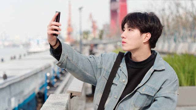 Portrait of a handsome Chinese young man in jeans holding his mobile phone to take pictures for himself, young people like selfie.