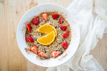 Superfood breakfast oatmeal bowl with fresh strawberries, orange slices, and maple syrup, on wooden table, linen napkin, modern tile, top view