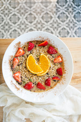 Superfood breakfast oatmeal bowl with fresh strawberries, orange slices, and maple syrup, on wooden table, linen napkin, modern tile, top view
