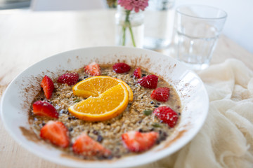 Superfood breakfast oatmeal bowl with fresh strawberries, orange slices, and maple syrup, flowers on table, wooden background