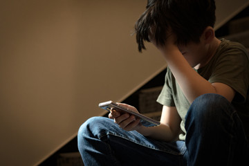 Cyber bullying concept. Young Asian preteen/teenage boy sitting at stair, covering his face with...