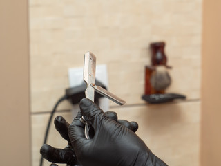 Stylist preparing open razor for shaving, close up view. Masters hands in black rubber gloves. Interior of barbershop. Selective soft focus. Blurred background