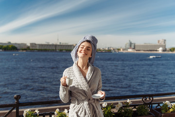 Young beautiful happy woman after a shower in a bathrobe and towel standing on the balcony with a cup of coffee. Cute sexy girl happy sunny morning on the ..background of the city.