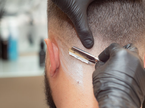 Hairstylist shaving clients neck with straight razor, close up view. Hands in black rubber gloves with open razor. Photographed in hairdressing saloon. Selective soft focus. Blurred background