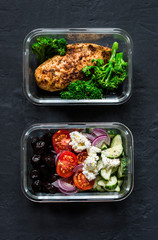 Two healthy balanced lunch boxes with greek salad, baked chicken breast and broccoli on a dark background, top view