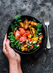 Smoked salmon, baked potatoes, fresh vegetables salad breakfast bowl in women's hands on a dark background, top view