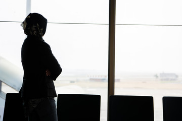 the silhouette of the rear view of a veiled Muslim woman standing in front of a large window in the airport waiting room