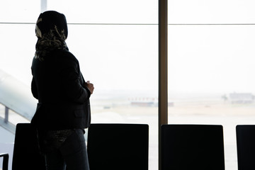 the silhouette of the rear view of a veiled Muslim woman standing in front of a large window in the airport waiting room