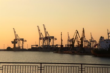Fototapeta na wymiar Cranes work with containers, in the commercial port of Odessa. In the biggest harbor of the Black sea. Photo taket during golden hours. Colorful sky and Black sea in the background.