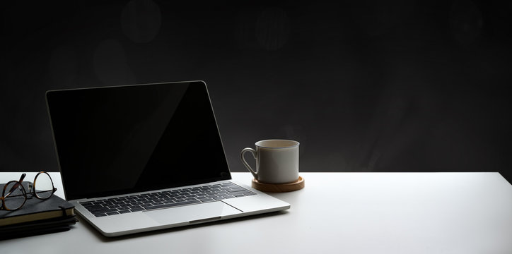Open laptop computer with coffee cup and notebook on white table and blank wall background