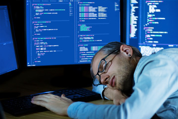 Senior software developer workplace. Programmer sleeping at work against displays with complex...