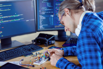Developer is connecting breadboard to microcontroller for creating prototype of electronic device. Chips, resistors, light-emitting diodes on desktop of hardware engineer. Program code on background. - obrazy, fototapety, plakaty