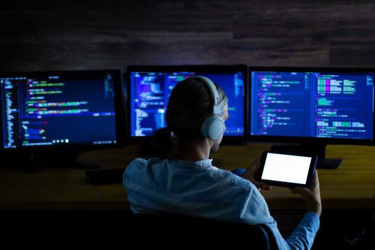 Software developer freelancer with white headphones and glasses working with program code: C++, Java, Javascript on wide displays and tablet at night. Develops new web, desktop, mobile application.