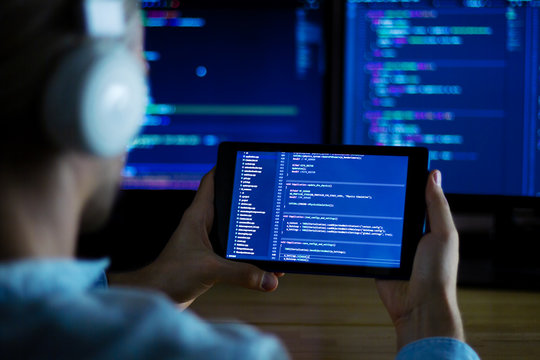 Software developer freelancer with white headphones and glasses working with program code: C++, Java, Javascript on wide displays and tablet at night. Develops new web, desktop, mobile application.
