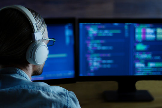Modern software developer freelancer with white headphones working with program code: C++, Java, Javascript on wide displays at night. Develops new web, desktop, mobile application or framework.