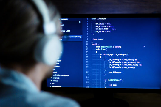 Smart software developer, freelancer with white headphones develops new web desktop application in workplace at night. Working with complex program code: c++, java, javascript on wide display.