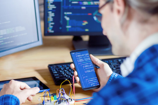Developer is connecting breadboard to microcontroller. Man is holding smartphone with program code software for controlling electronic device. Chips, resistors, diodes on desktop of hardware engineer.