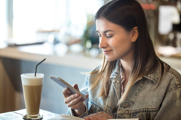 Young girl sitting in a cafe drinking coffee and looking at the phone