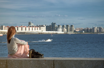 A young girl sitting on a granite parapet on the shore of the Gulf of Finland in St. Petersburg on a blurred background of a bay with modern buildings.