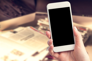 Hand holding a smartphone with a blank screen. Newspapers blurred in the background. Concept of the morning press and telephone in hands.