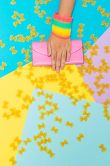 Hand with rainbow accessories holds pink purse or wallet among pasta bows on geometric background