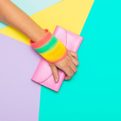 Hand with rainbow accessories holds pink purse on geometric background