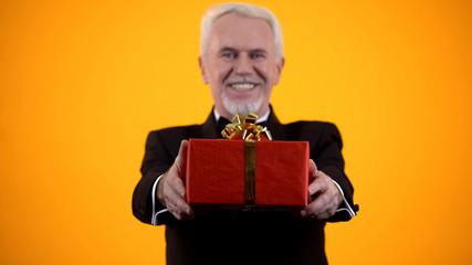 Cheerful male retiree in suit presenting red giftbox on camera, birthday present