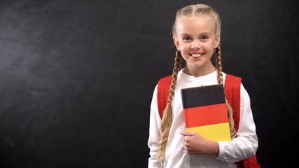 Smiling female pupil holding German language textbook, blackboard background