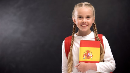 Smiling schoolgirl holding Spanish language book against blackboard background