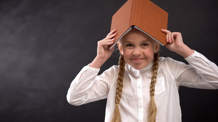 Excited female pupil with book on head smiling on camera, education concept