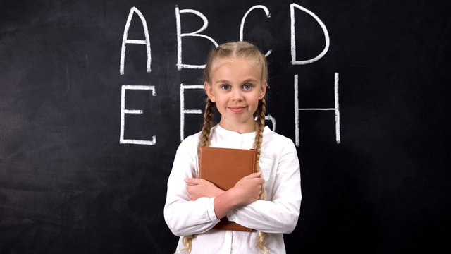 Pretty elementary school pupil standing against alphabet written on blackboard
