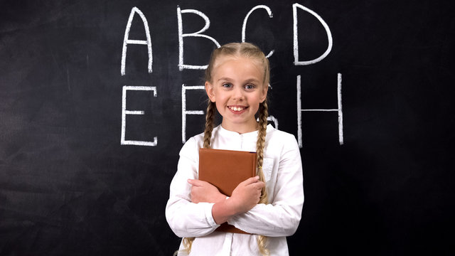 Smiling schoolgirl hugging book, alphabet written on blackboard behind, system