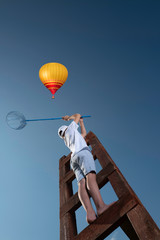 A little boy on a wooden staircase leading to the sky with butterfly net wants to catch something interesting 