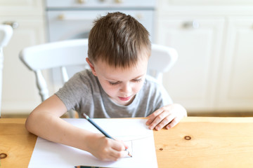 Little boy sits at a table in a bright kitchen and draws with pencils
