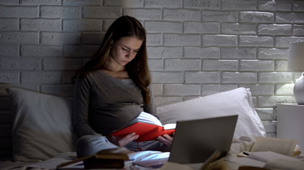 Pregnant woman reading book in front of laptop, worrying about childbirth