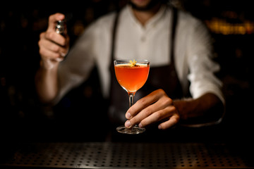 Male bartender serving a cocktail in the glass with a caramelized lemon slice and a little yellow flower spraying on it