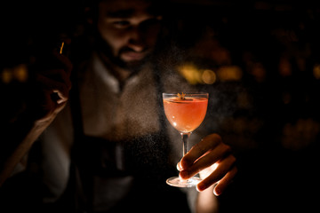 Attractive bartender serving a brown orange cocktail spraying on it in the dark