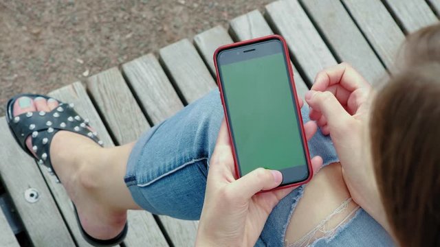 Young beautiful girl in the park. Holds in his hands a black smartphone with a green screen. Female hand holds a phone with chrome kay. Makes the zoom with two fingers, examines the map.