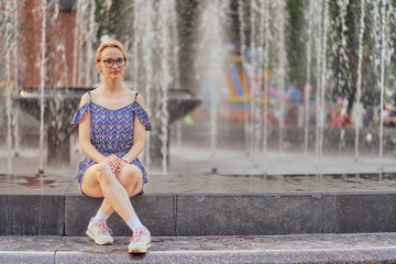 young athletic girl with glasses in the park. sits by the fountain in a short dress and sneakers